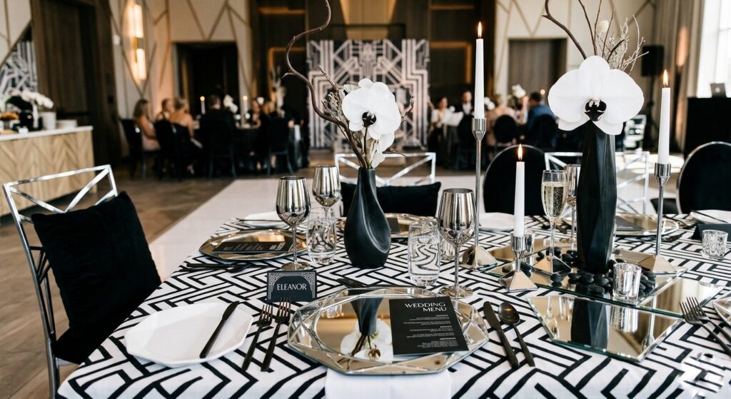 Modern elegant wedding tablescape with geometric mirrored charger plates, chrome candlestick holders, graphic black and white linen, and a sculptural single-stem white floral arrangement creating a high-contrast Art Deco aesthetic