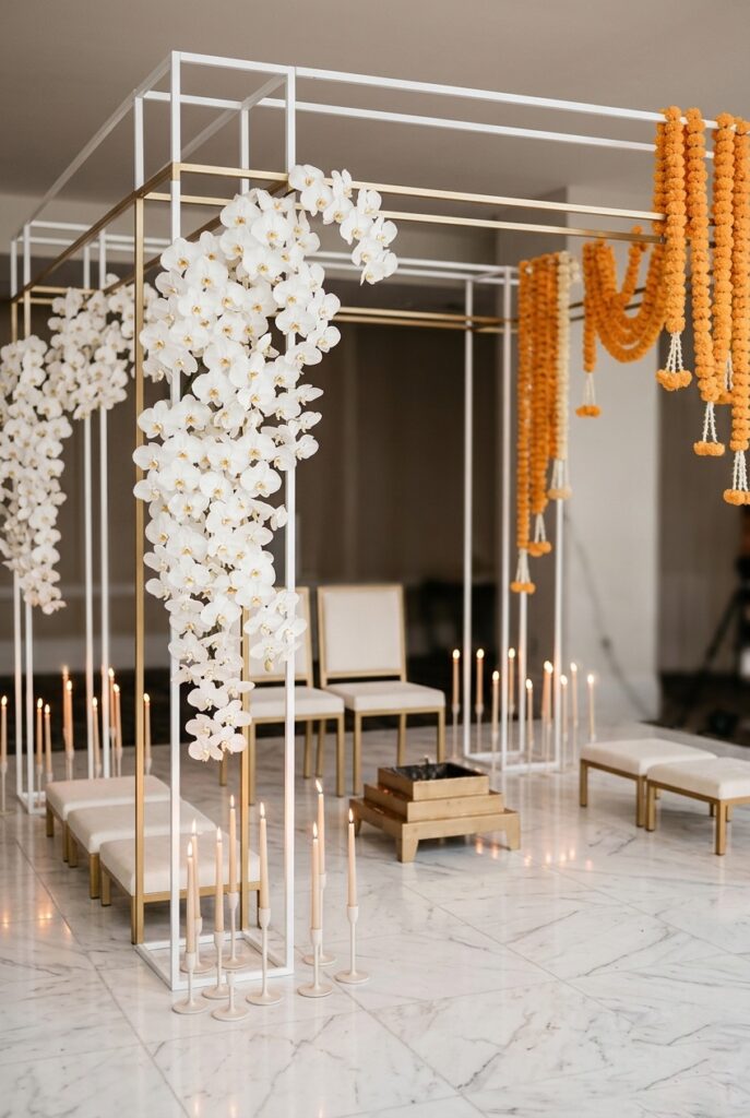 Minimalist modern Indian wedding mandap with a white and gold geometric frame, cascading white orchids, soft marigold garland accents, and warm candlelight on a polished marble floor