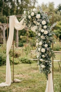 Simple DIY wedding ceremony arch made from a metal frame decorated with eucalyptus bundles, white roses, and baby's breath from a grocery store, standing outdoors in natural light