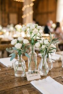 White spray roses and eucalyptus stems in clear glass bud vases on a rustic wood wedding table — portrait orientation