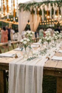 Neutral cheesecloth table runner draped loosely over a long farm table at an outdoor wedding reception — portrait orientation
