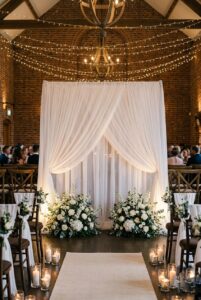 Simple white sheer fabric wedding ceremony backdrop with two white floral arrangements at base in an indoor venue — portrait orientation