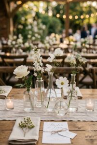 Cluster of five mismatched clear glass bud vases with single stem white flowers on a linen wedding table — portrait orientation