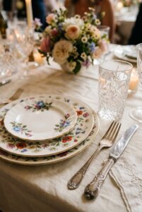 Mismatched genuine vintage china plates, crystal stemware, and antique silverware laid on a linen tablecloth at a wedding reception — portrait 2:3