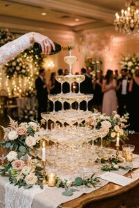 Elegant vintage wedding champagne tower with stacked coupe glasses and champagne being poured from the top — portrait 2:3