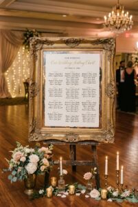 Large ornate gold antique frame propped on a vintage easel displaying a wedding seating chart with calligraphy lettering — portrait 2:3