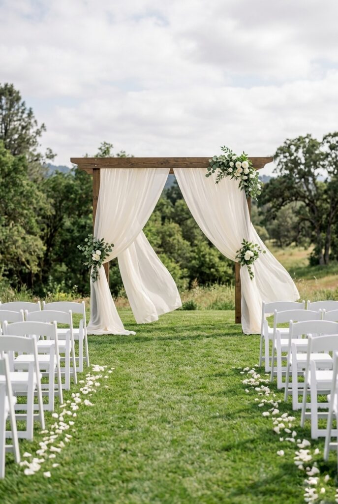 Sheer ivory chiffon curtain panels draped from a simple wooden arch creating a dreamy wedding ceremony backdrop — portrait format