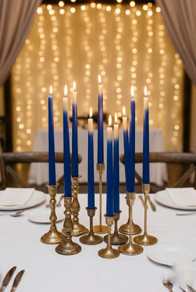 Close-up of a wedding table with clusters of tall blue taper candles in mismatched brass and gold candleholders at varying heights, no flowers, on a white linen tablecloth — the blue wax softening to cream at the flame