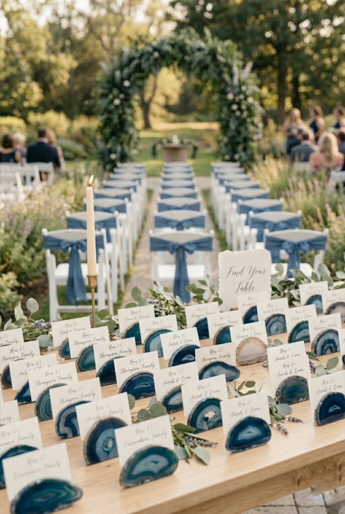 Wedding escort card display table with blue-toned natural agate slices used as individual card holders, each slice showing translucent blue and white mineral bands, arranged on a light wood surface with a small greenery sprig and a single candle