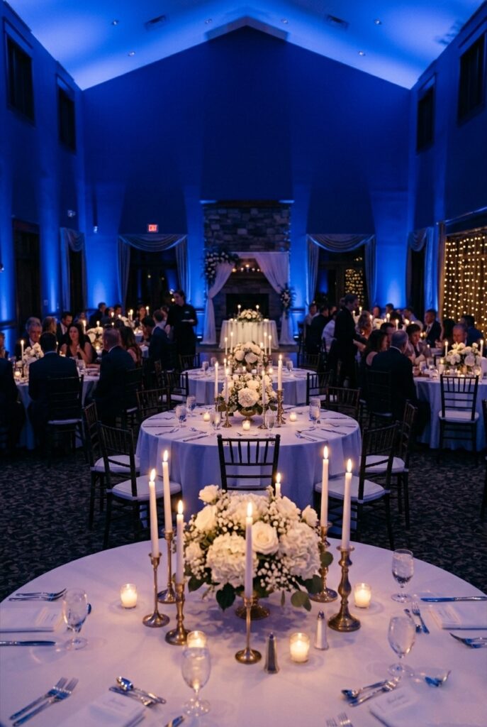 Indoor wedding reception venue with deep sapphire blue uplighting washing the walls, creating a dramatic and moody atmosphere over round tables with white linens, white floral centerpieces, and warm candlelight on the tables — the blue walls contrasting with the warm table lighting