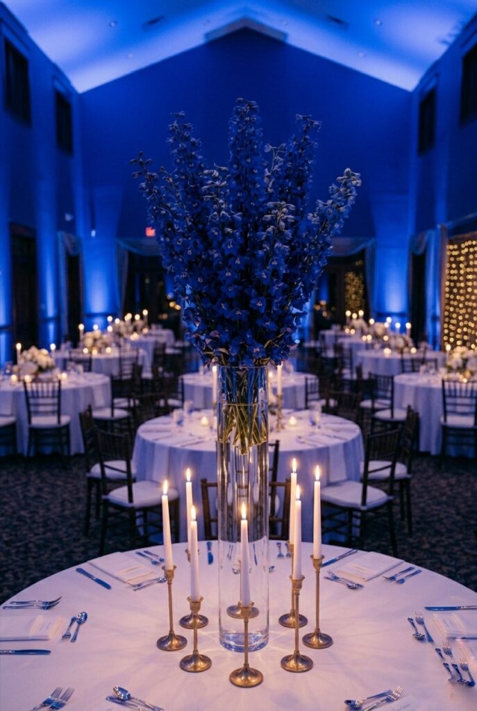 Tall wedding table centerpiece with a single variety of deep blue delphiniums arranged in a clear glass cylinder vase — no filler flowers, no greenery, no mixed varieties — at dramatic height against a white tablecloth and simple gold candleholders