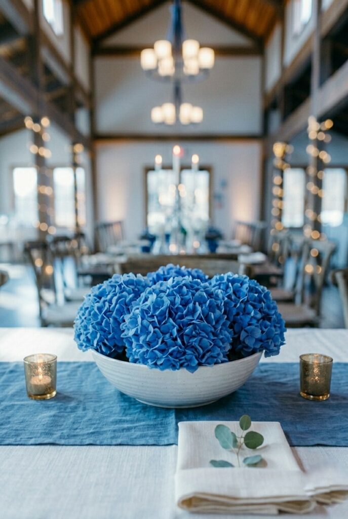 IMAGE 5 HERE Alt text: Three true-blue hydrangea heads in a wide white ceramic bowl centerpiece on a dusty blue linen runner at a wedding table Image prompt: Photorealistic portrait shot of a wedding table centerpiece. A wide shallow white ceramic bowl holds three full Nikko Blue hydrangea heads in a true medium blue, packed closely together so the blooms form one continuous mass of color. The bowl sits on a dusty blue washed-linen runner. Two small brass votive candle holders with lit tea lights are placed at opposite sides of the bowl. A folded ivory napkin with a small sprig of eucalyptus is visible at the frame's lower right. Soft natural window light from the upper left emphasizing the true blue hydrangea color. No text overlays. Generate in vertical portrait orientation, 2:3 aspect ratio, optimized for mobile display.