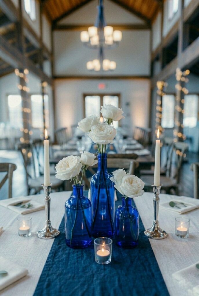 IMAGE 2 HERE Alt text: Three vintage cobalt blue glass bottles with white garden roses as a wedding table centerpiece on a linen runner Image prompt: Photorealistic portrait shot of a blue wedding table centerpiece. Three vintage cobalt blue glass bottles of graduated heights — one tall narrow apothecary bottle approximately 10 inches, one medium round-body bottle approximately 7 inches, one short squat bitters bottle approximately 5 inches — each holding two to three stems of white garden roses in slightly different stages of bloom. The bottles are grouped in an asymmetric cluster on a white linen runner. A small lit clear glass votive sits at the base of the cluster. Soft natural window light from the left creating a faint cobalt glow through the glass. No text overlays. Generate in vertical portrait orientation, 2:3 aspect ratio, optimized for mobile display.