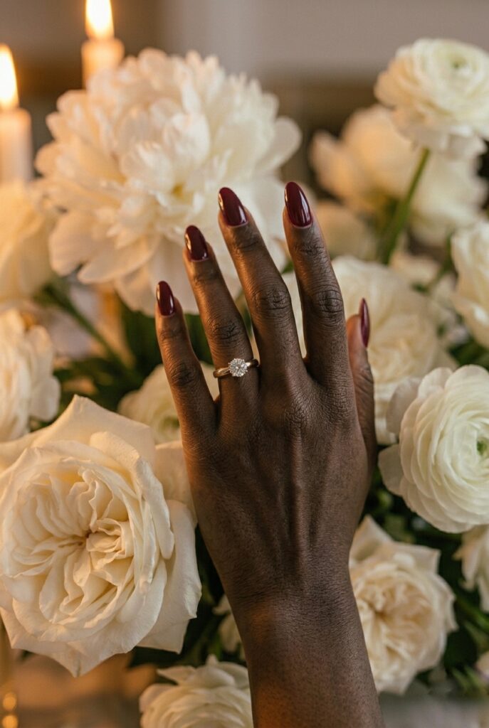 IMAGE 7 HERE Alt text: Deep-toned Black bride wearing a rich burgundy wine nail polish on long almond nails, hand resting on white bridal bouquet of peonies and garden roses Image prompt: Photorealistic close-up of a deep-toned hand with rich ebony complexion wearing a deep burgundy wine gel manicure on long almond-shaped nails, hand resting on a lush white bridal bouquet of peonies and garden roses, warm candlelit atmosphere with amber light accenting the skin tone and nail color, rich contrast between dark nails and white blooms, editorial bridal photography, no text overlays. Generate in vertical portrait orientation, 2:3 aspect ratio, optimized for mobile display.