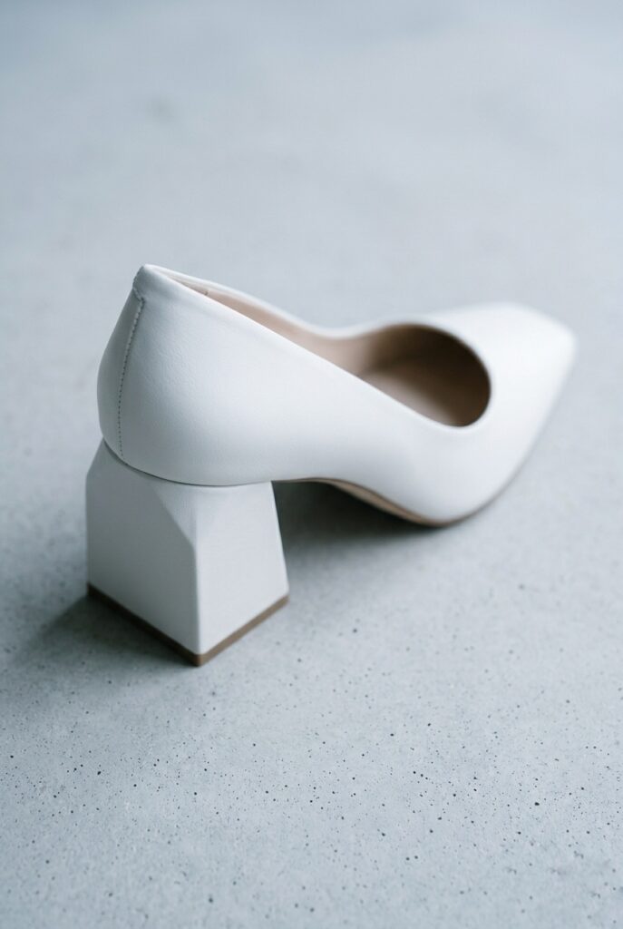 IMAGE 2 HERE Alt text: White sculptural block heel bridal shoe with geometric heel architecture on a concrete surface, clean cool natural daylight Image prompt: Photorealistic close-up portrait of an architectural white leather bridal block heel shoe with a geometric squared-off heel that has a subtle sculptural indentation. Pointed square toe, clean minimal upper with no embellishment. Placed on a smooth pale concrete surface. Cool clean natural daylight from above, no warm tones. The focus is on the heel's architectural geometry. Background is blurred pale concrete. No text overlays. Generate in vertical portrait orientation, 2:3 aspect ratio, optimized for mobile display.