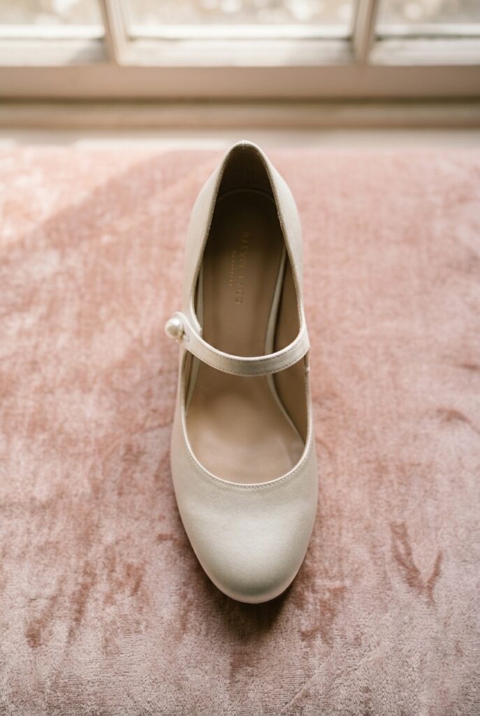 Photorealistic close-up portrait of an ivory satin Mary Jane bridal heel with a single center bar strap with a small pearl button closure, rounded toe, and a 2.5-inch block heel. Placed on a pale blush pink velvet fabric surface. Soft warm natural window light from above illuminates the satin and the pearl button detail. Background is softly blurred pink velvet. No text overlays. Generate in vertical portrait orientation, 2:3 aspect ratio, optimized for mobile display.