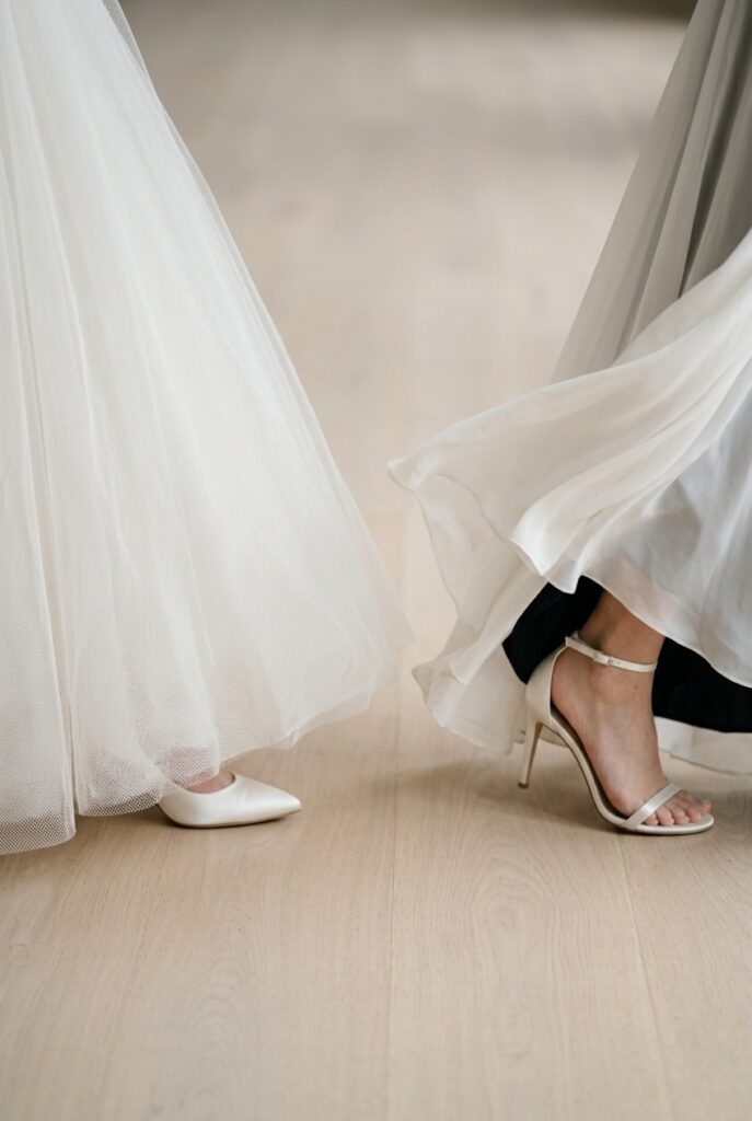 Image prompt: Photorealistic close-up portrait on pale smooth wood floor. Two gown hem cross-sections shown side by side for comparison. Left: a heavyweight structured ivory tulle ballgown hem at mid-stride, the dense layers of netting lifting slightly from the floor, showing only the very toe of an ivory pump beneath. Right: a lightweight ivory chiffon hem in mid-stride, the fabric billowing upward and showing the complete shoe and lower ankle clearly. Soft natural window light from above illuminates both hems equally. Background is blurred pale wood floor. No text overlays. Generate in vertical portrait orientation, 2:3 aspect ratio, optimized for mobile display.