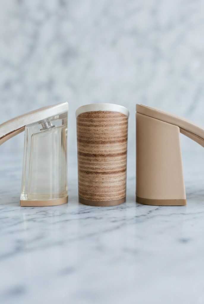 IMAGE 9 HERE Alt text: Three chunky bridal heel columns shown in cross-section or profile comparison — lucite core, wood core, foam core — on pale marble with clean natural light showing material differences Image prompt: Photorealistic close-up portrait on pale cool marble. Three chunky heel columns from ivory bridal pumps shown side by side in close profile, each representing a different core material. Left: a translucent lucite-core chunky heel where the pale resin interior is faintly visible through a thin ivory satin covering. Center: a stacked wood-core heel with visible natural wood grain at the exposed heel edge. Right: a foam-core heel with a smooth uniform surface and no visible grain or texture at the edge. Clean cool natural overhead daylight from above creates even illumination showing the material differences at each heel edge. Background is blurred pale marble. No text overlays. Generate in vertical portrait orientation, 2:3 aspect ratio, optimized for mobile display.