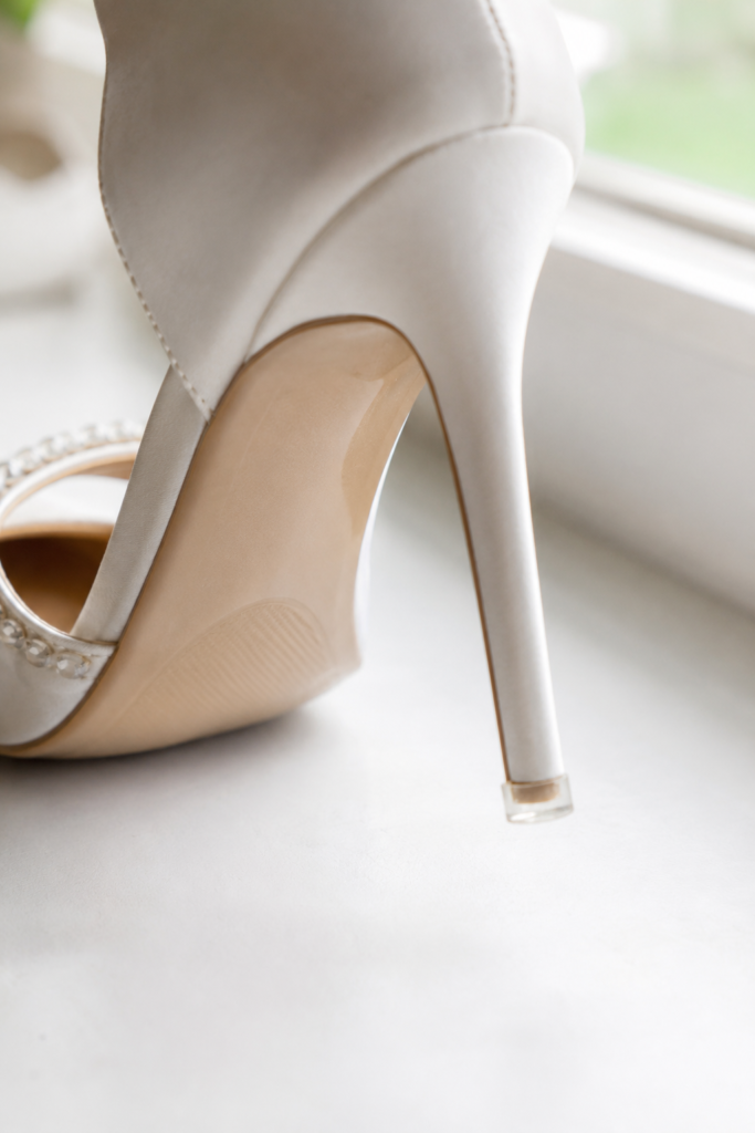 IMAGE 10 HERE Alt text: Bride's bridal heel shoes protected with heel cap and sole protector before outdoor wedding ceremony Image prompt: Photorealistic close-up image of an ivory bridal heel shoe on a white surface, with a clear rubber heel cap applied to the slim heel tip, visible as a small transparent disk at the base of the heel. Soft natural window light from above. No text overlays. Detail-focused editorial product shot. Generate in vertical portrait orientation, 2:3 aspect ratio, optimized for mobile display.