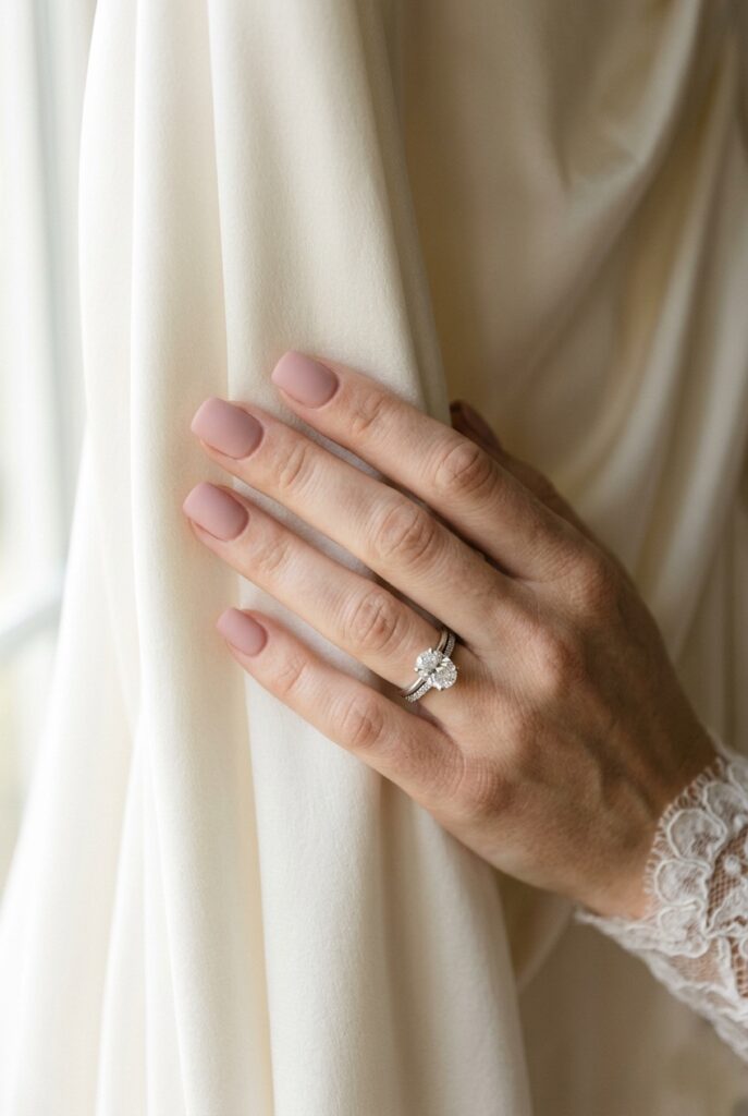 Photorealistic close-up of a bride's hand with short square-shaped nails finished in a soft matte blush pink — a dusty rose tone with no shine. Hand resting against silk ivory fabric. Soft natural window light, understated and minimalist composition. Engagement ring visible. No text overlays. Generate in vertical portrait orientation, 2:3 aspect ratio, optimized for mobile display.